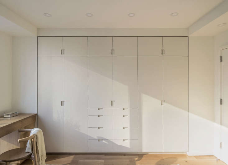 In a Workstead-designed bedroom, built-in closets feature painted millwork with flush pivot doors and routed pulls. The storage wall is fitted with closets, drawers, overhead cupboards, and, in the center, a cabinet that holds the TV. Photograph by Matthew Williams, courtesy of Workstead, from The Artful Shoebox Apartment, Workstead Edition.